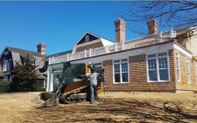Man operating a skid steer next to a house with cedar shingle siding on a sunny day.