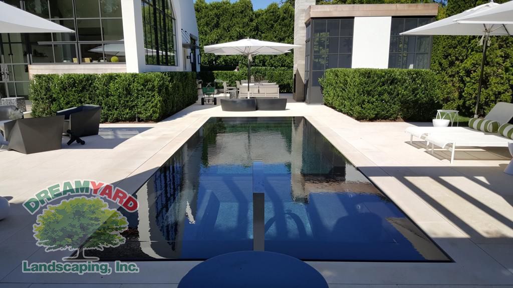 Luxury backyard pool with white patio, green hedges, and outdoor furniture.
