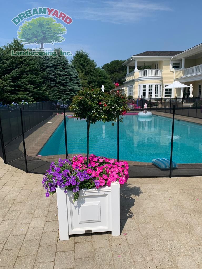 White planter box with purple and pink flowers, trees, pool, and a large yellow house.