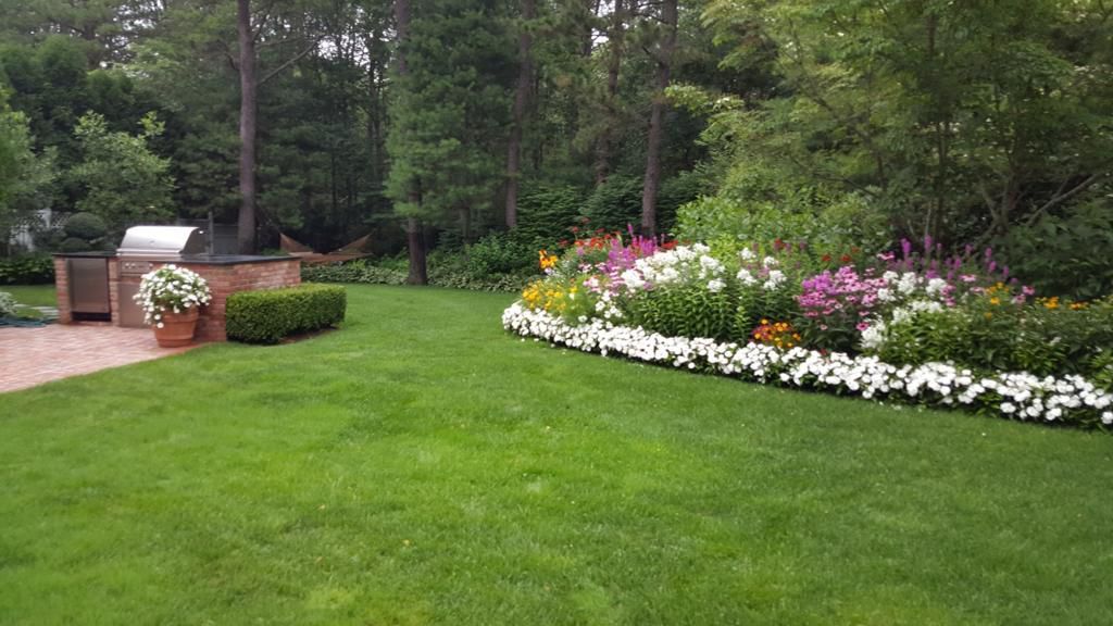 Lush green backyard with flowering garden bed, built-in grill, and trees.