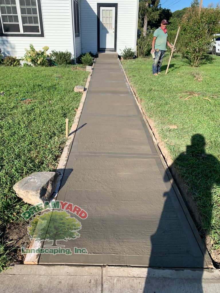 New concrete walkway being installed at a white house. Person in yard, wooden forms on sides.