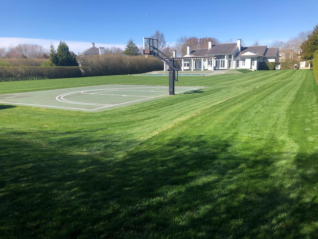 Green lawn with basketball court and large white house on a sunny day.