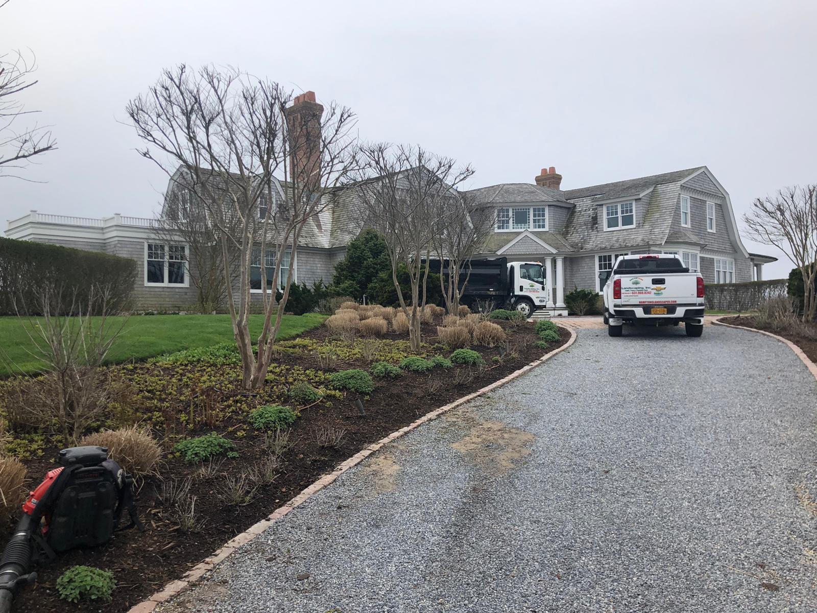 A large gray house with a gravel driveway and landscaping. A white truck and small delivery truck are parked in front.