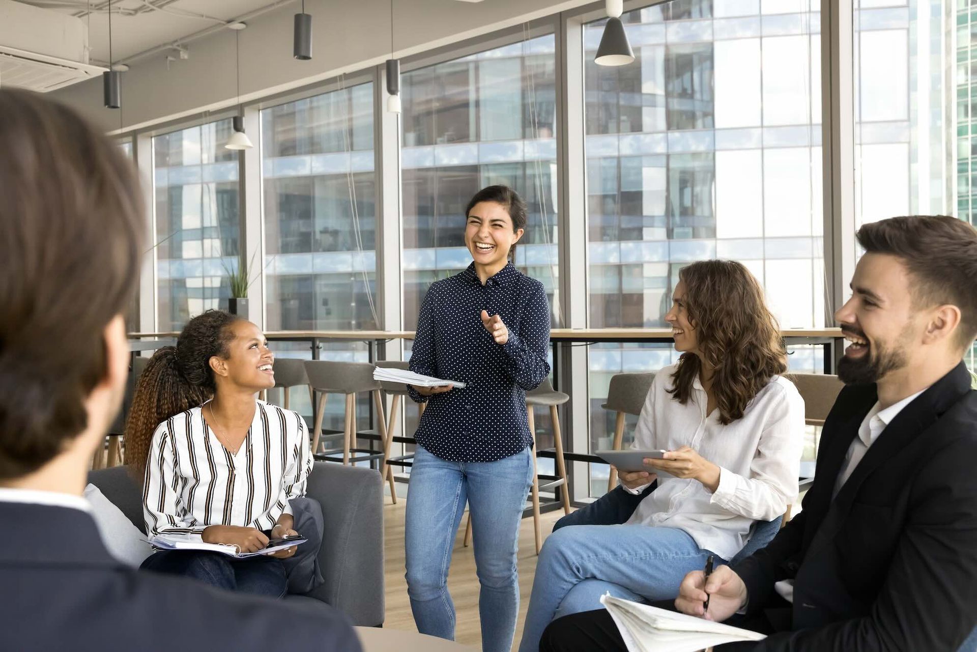 A woman leading a team meeting in a modern office; smiling, gesturing, holding papers.