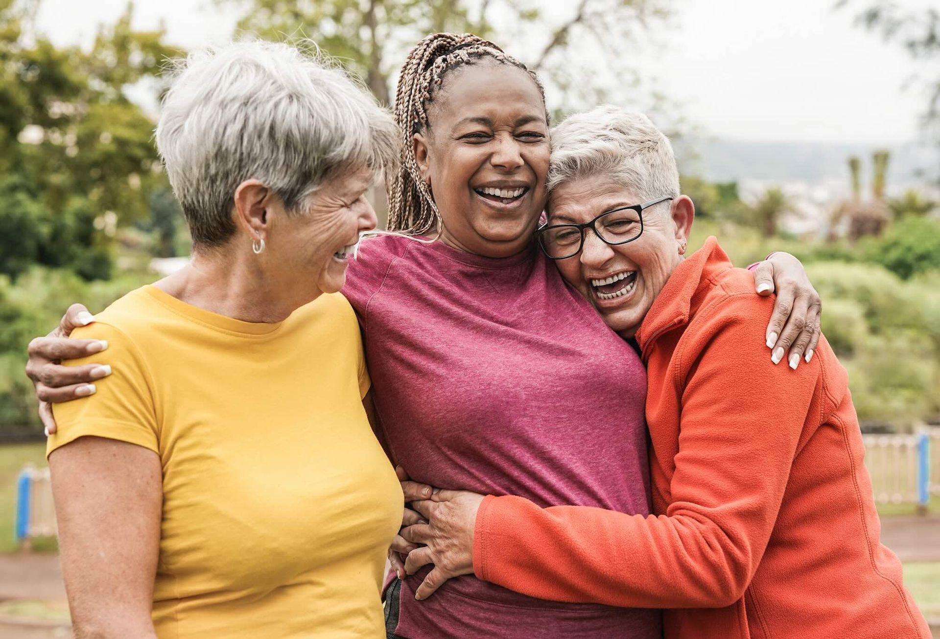 Three smiling women hugging outdoors; one in yellow, one in red, and one in burgundy.