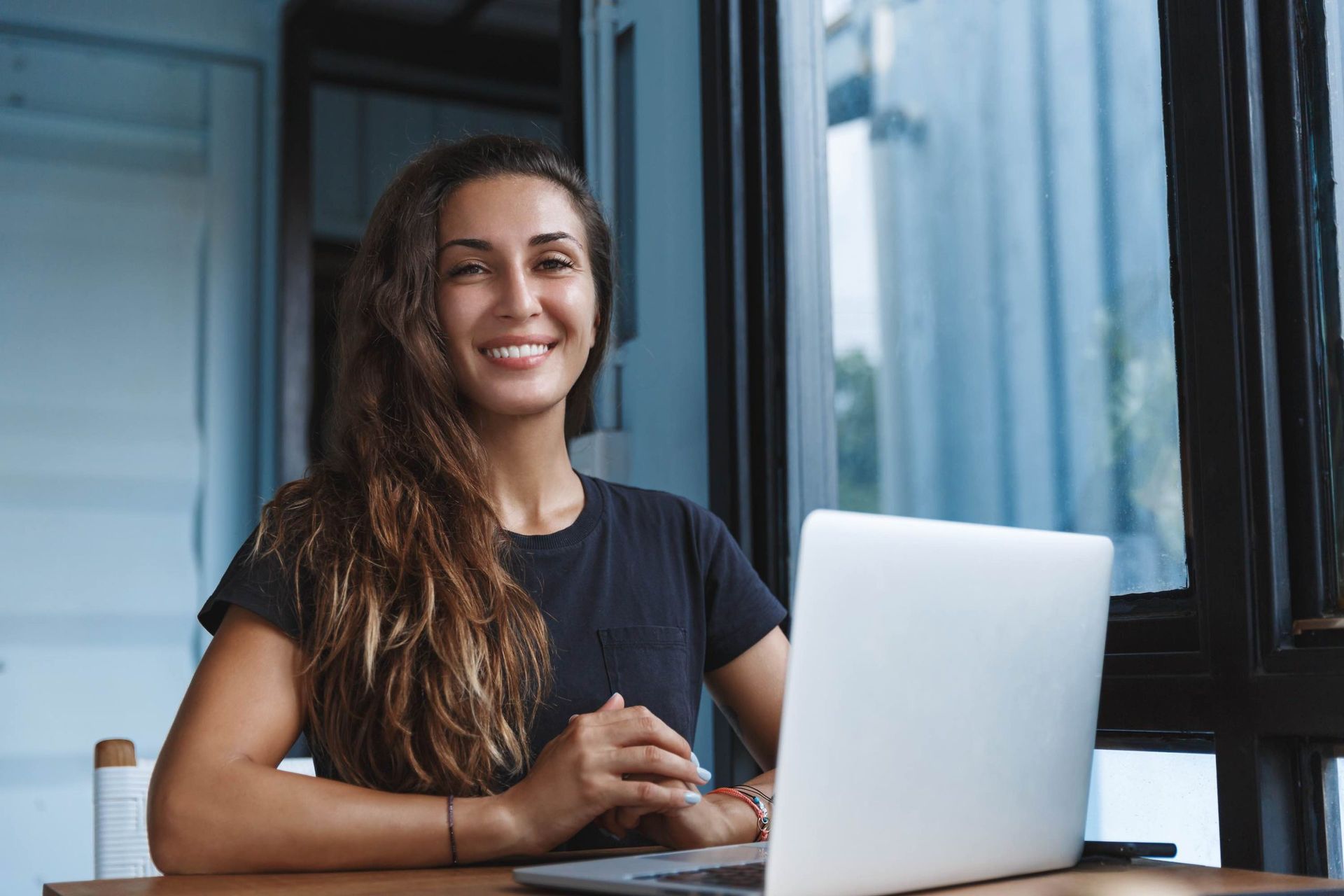 Woman with long brown hair smiling, sitting at a table with a laptop, near a window.
