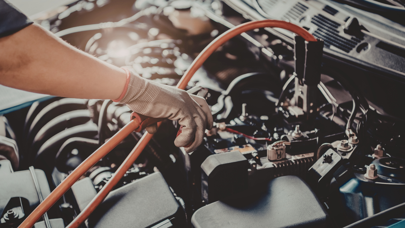 Gloved hand working on a car engine with orange cables in a garage