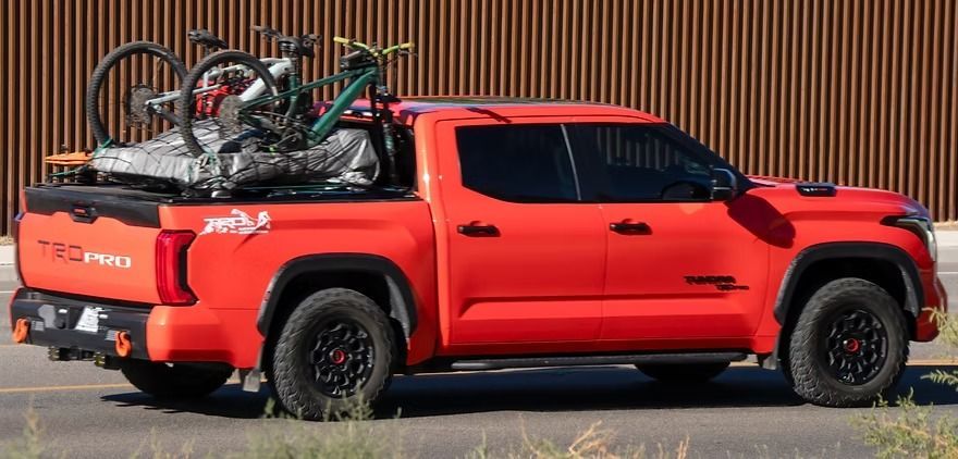 Red pickup truck with several bicycles secured in the bed, parked beside a corrugated metal wall.