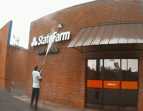 A person in a high-visibility vest uses a pressure washer to clean the concrete walkway outside a brick office building.