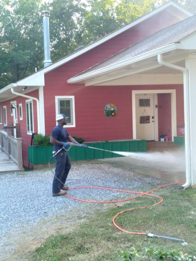A person in work clothes pressure-washes a red house with white trim on a sunny day.