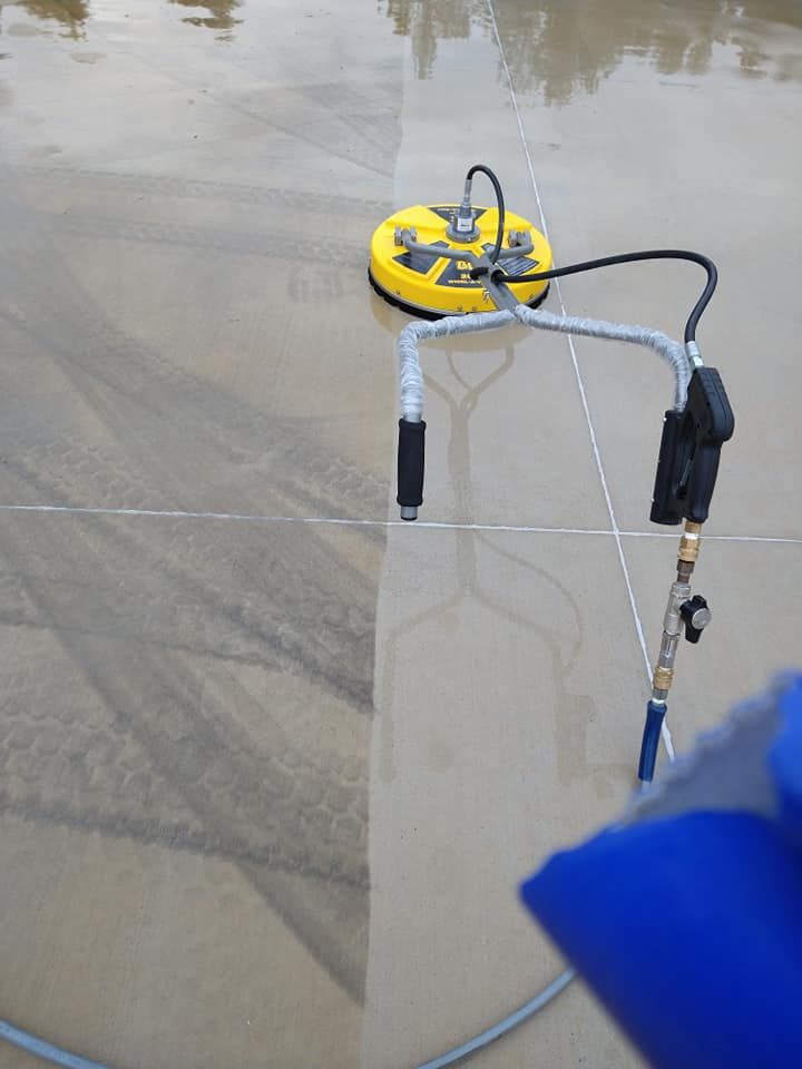 A yellow circular surface cleaner sits on a concrete driveway, showing a clear contrast between dirty and cleaned areas.