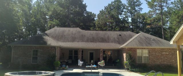 A backyard view of a brick house with a dark shingled roof, a covered patio, and a swimming pool in the foreground.