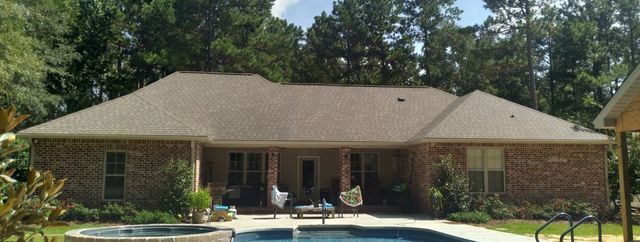 A brick house with a brown shingled roof, featuring a covered patio overlooking a swimming pool and wooded backyard.