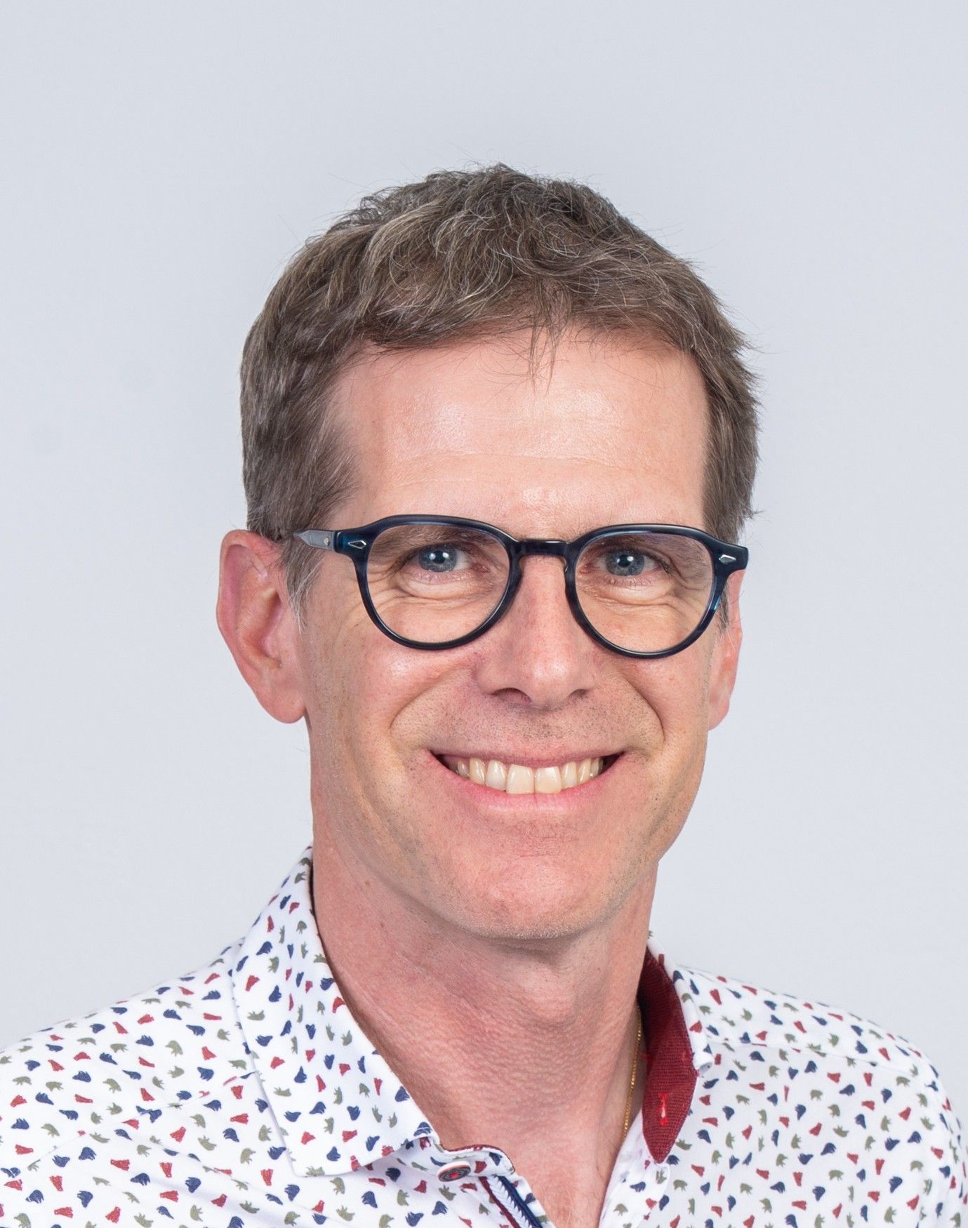 A smiling individual with short brown hair and dark-rimmed glasses, wearing a white shirt with a small, colorful pattern.