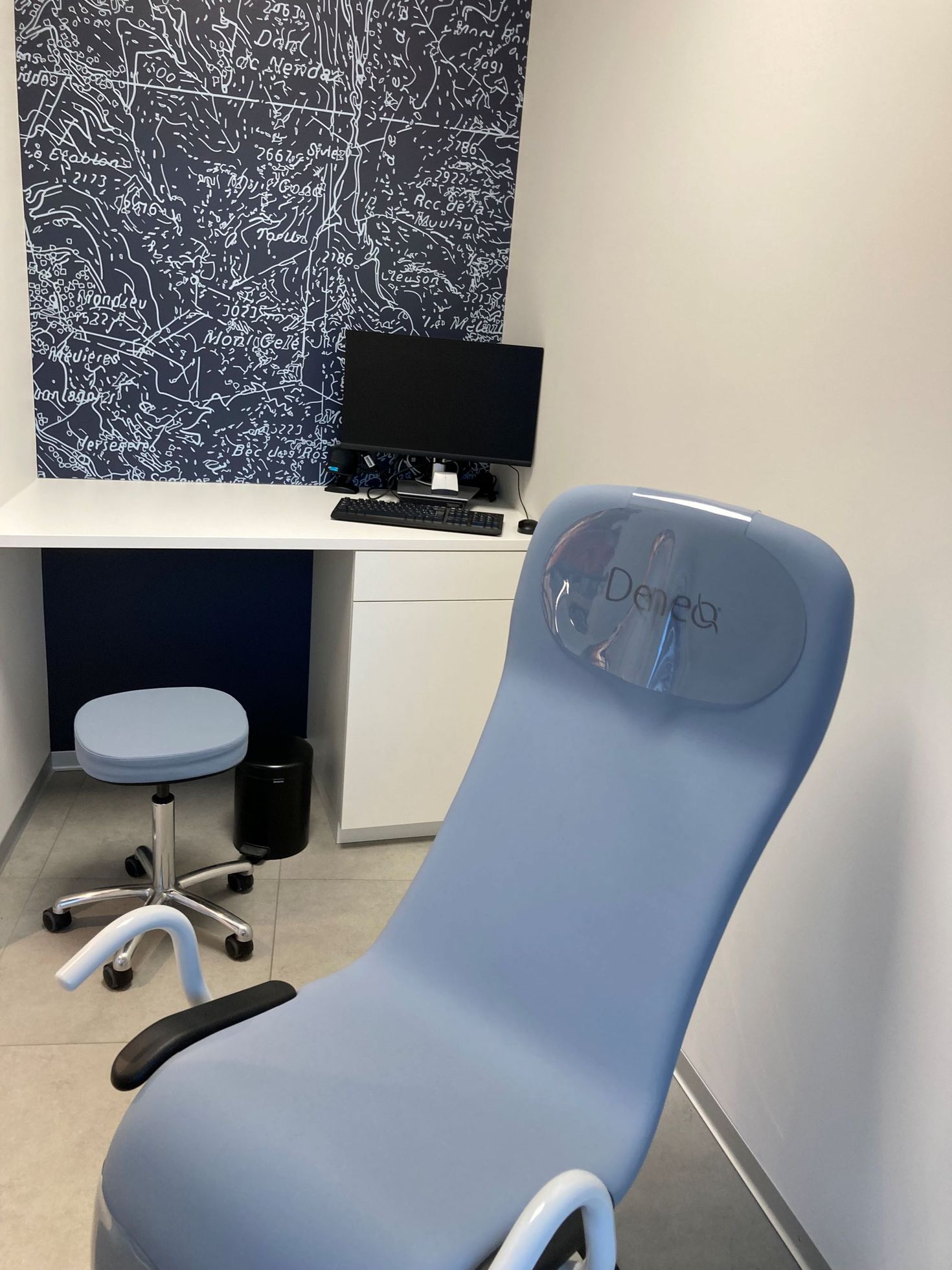 Ein hellblauer Untersuchungsstuhl steht vor einem weissen Schreibtisch mit einem Computermonitor an einer gemusterten Wand.