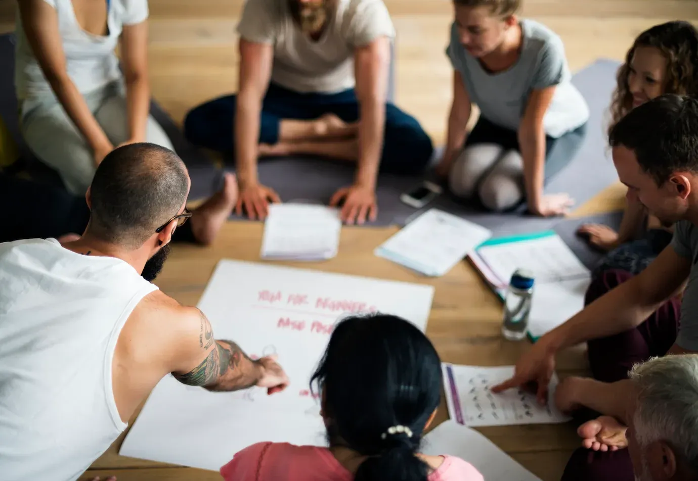 un grupo de personas están sentadas en el suelo en un círculo y miran un cartel que dice 