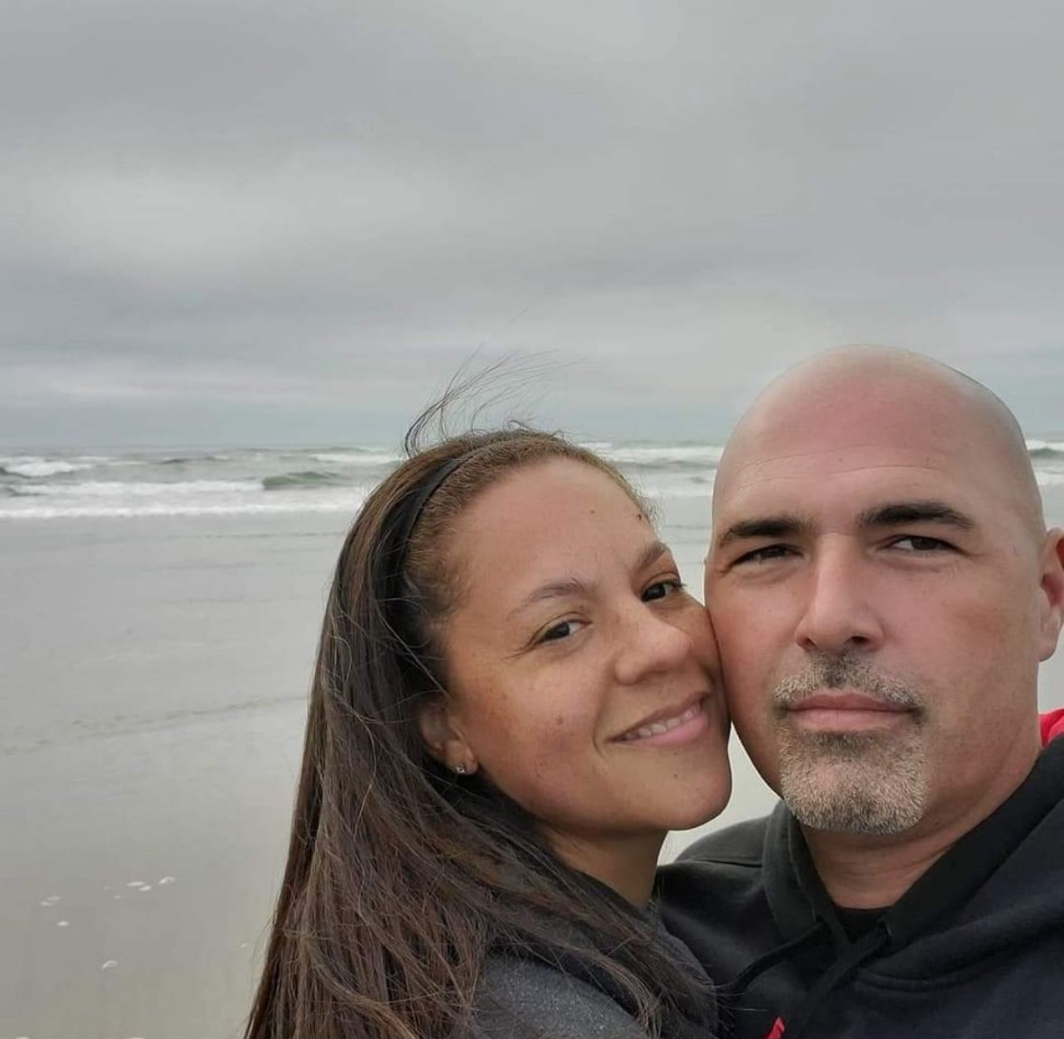 A couple posing for a selfie on a beach with an overcast sky and ocean waves in the background.