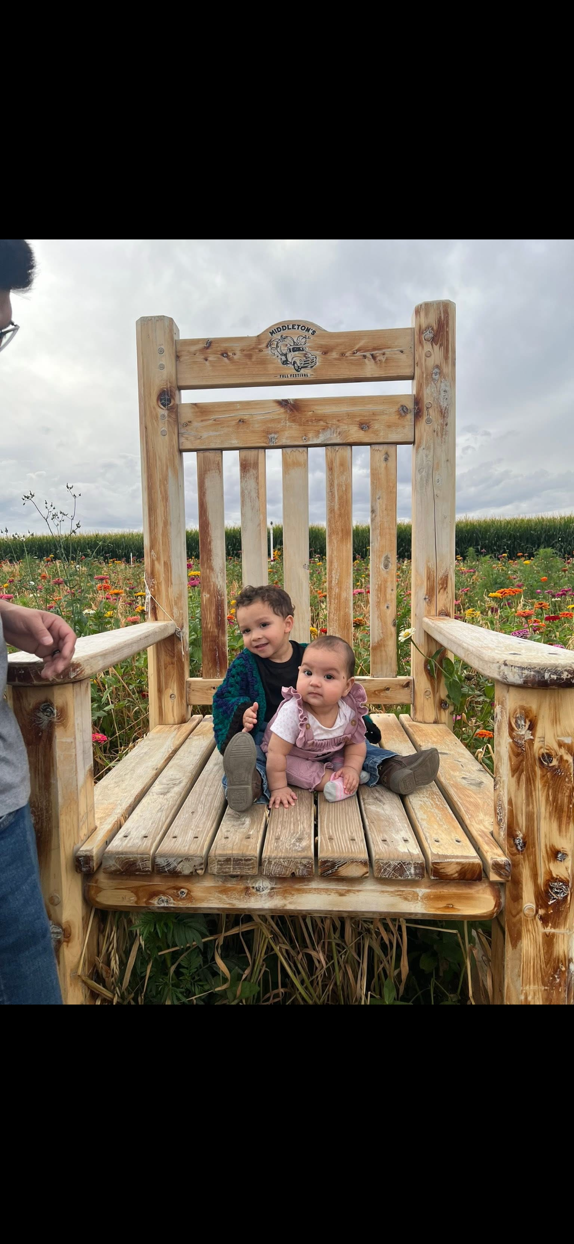 Two children sit on a large, rustic wooden chair outdoors in a field of flowers on a cloudy day.