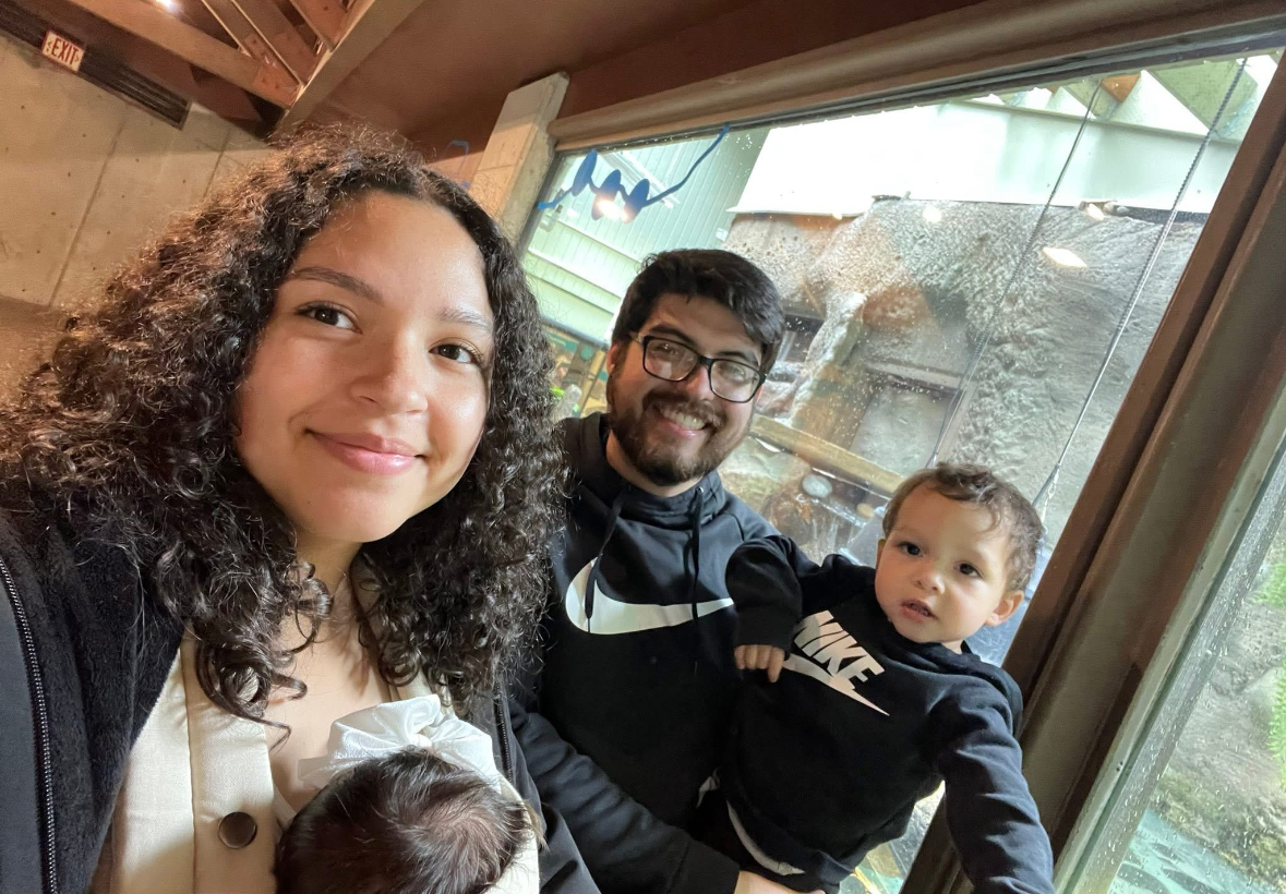A family of three smiles while posing for a selfie in front of a window at an indoor zoo or aquarium exhibit.