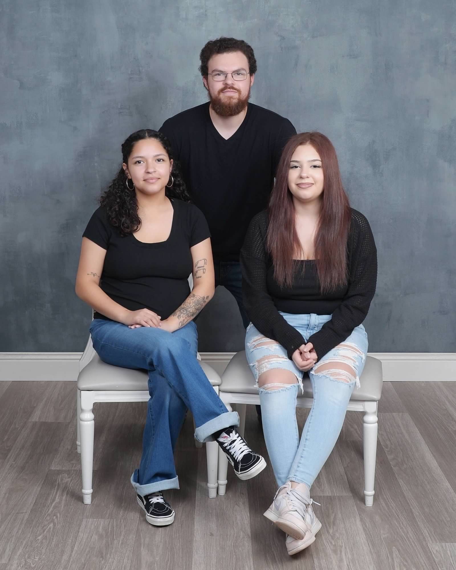Three people pose in front of a mottled blue backdrop; one man stands behind two seated people, all dressed in black tops.