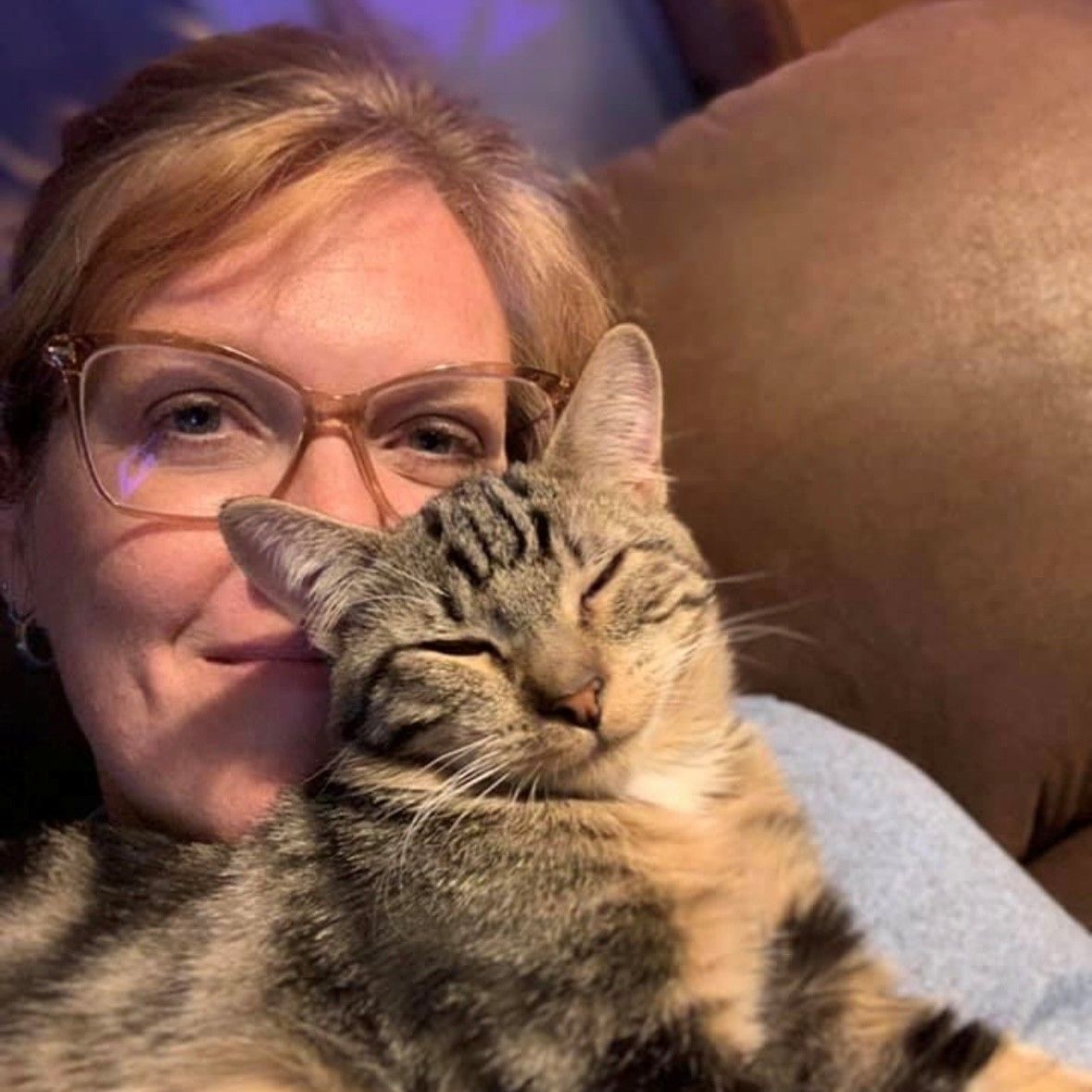 A woman with glasses smiles while resting her cheek against a sleeping brown tabby cat.