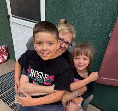 A smiling adult hugs two children while sitting on a porch in front of a dark green wall and a white door.