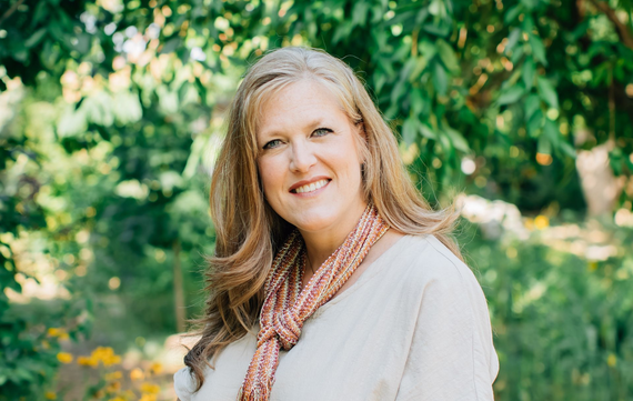 A smiling woman with long, light-brown hair and a patterned scarf standing in a lush, green, sunlit garden.