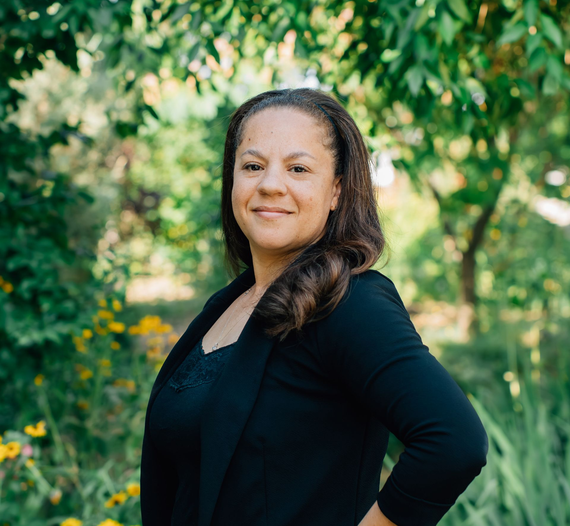 A person in a black blazer smiling softly, standing outdoors in a lush, green garden with yellow flowers.