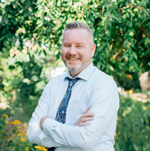 A person with a beard, wearing a light blue dress shirt and patterned tie, stands smiling with arms crossed in a garden.