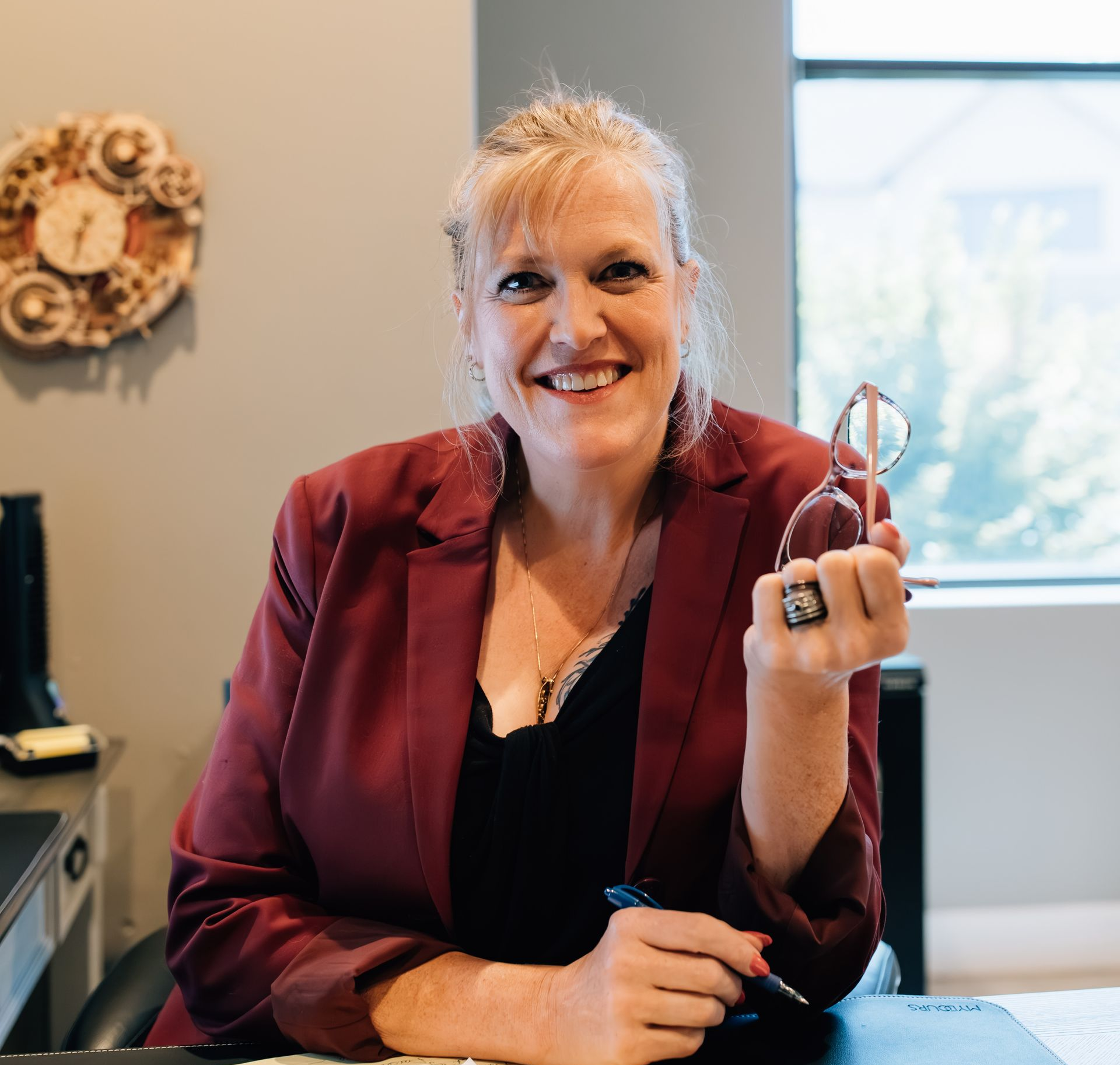 A professional smiling at a desk, wearing a maroon blazer and holding glasses and a pen in an office setting.