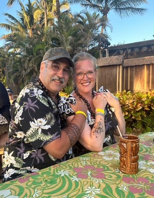 A couple smiling while sitting at a table with a tiki-themed cup, set against a backdrop of palm trees and tropical huts.