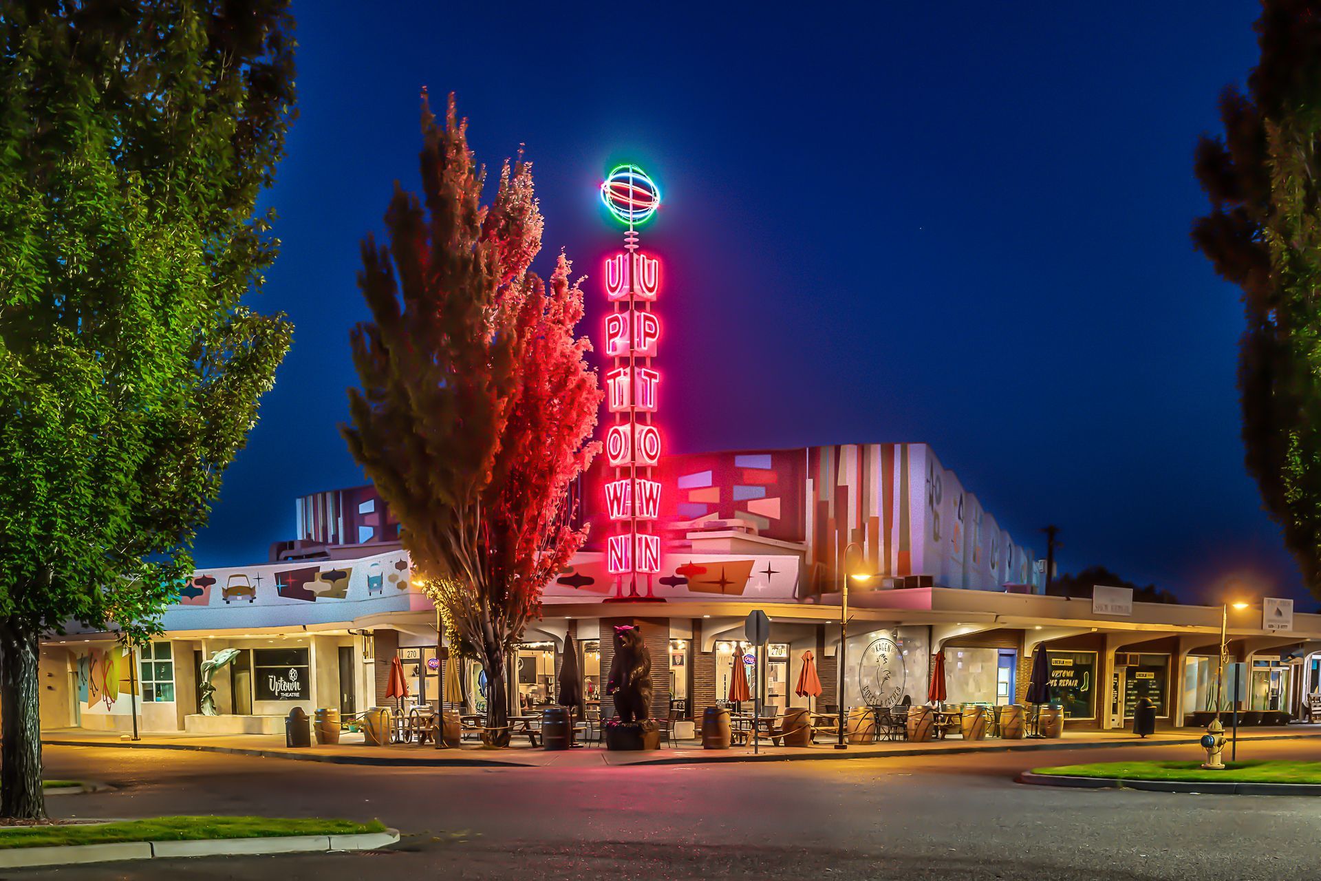 A neon-lit theater sign stands above a restaurant patio at dusk, surrounded by trees under a dark blue sky.