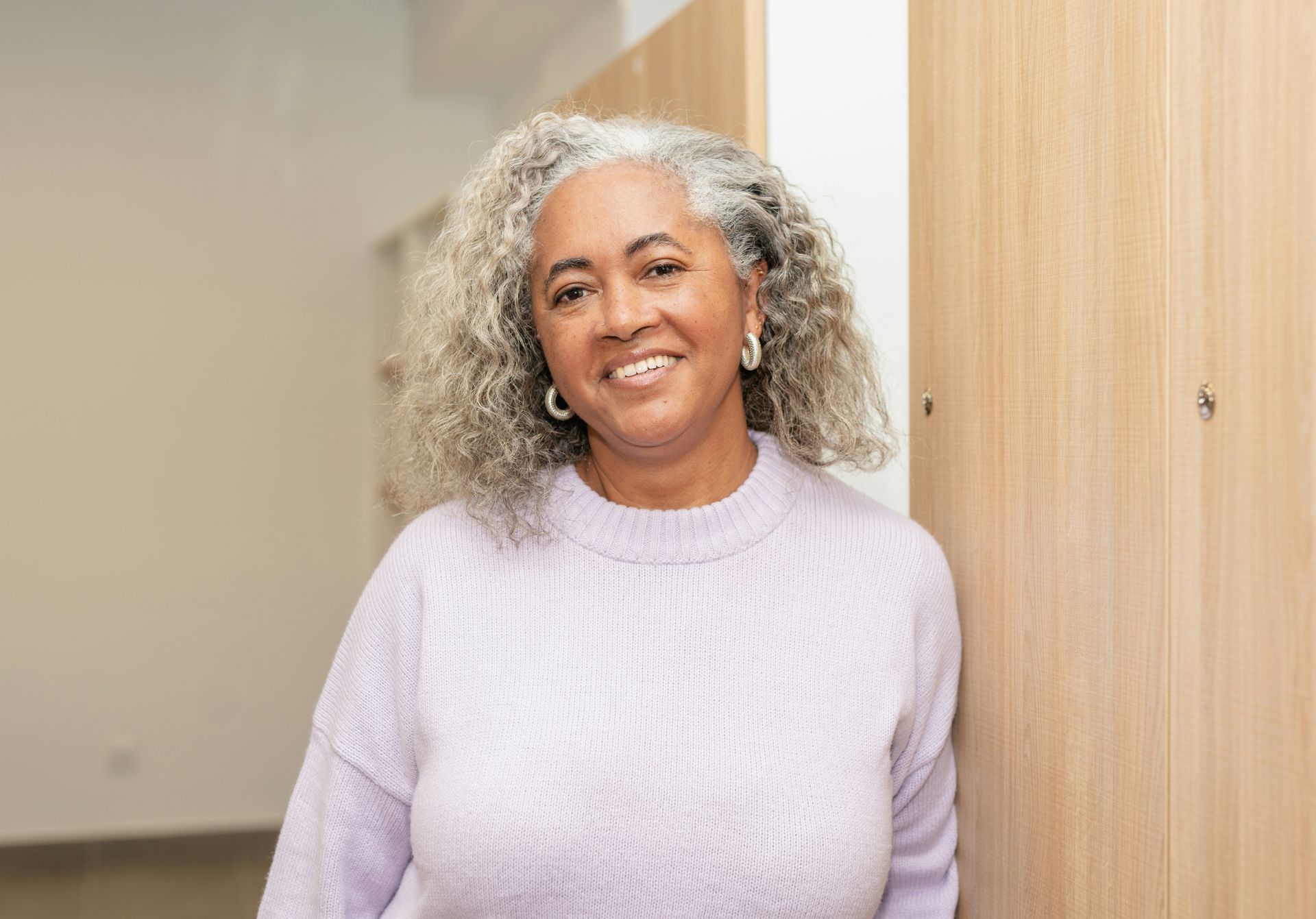 Smiling person with curly gray hair wearing a light purple sweater, leaning against light wood lockers.