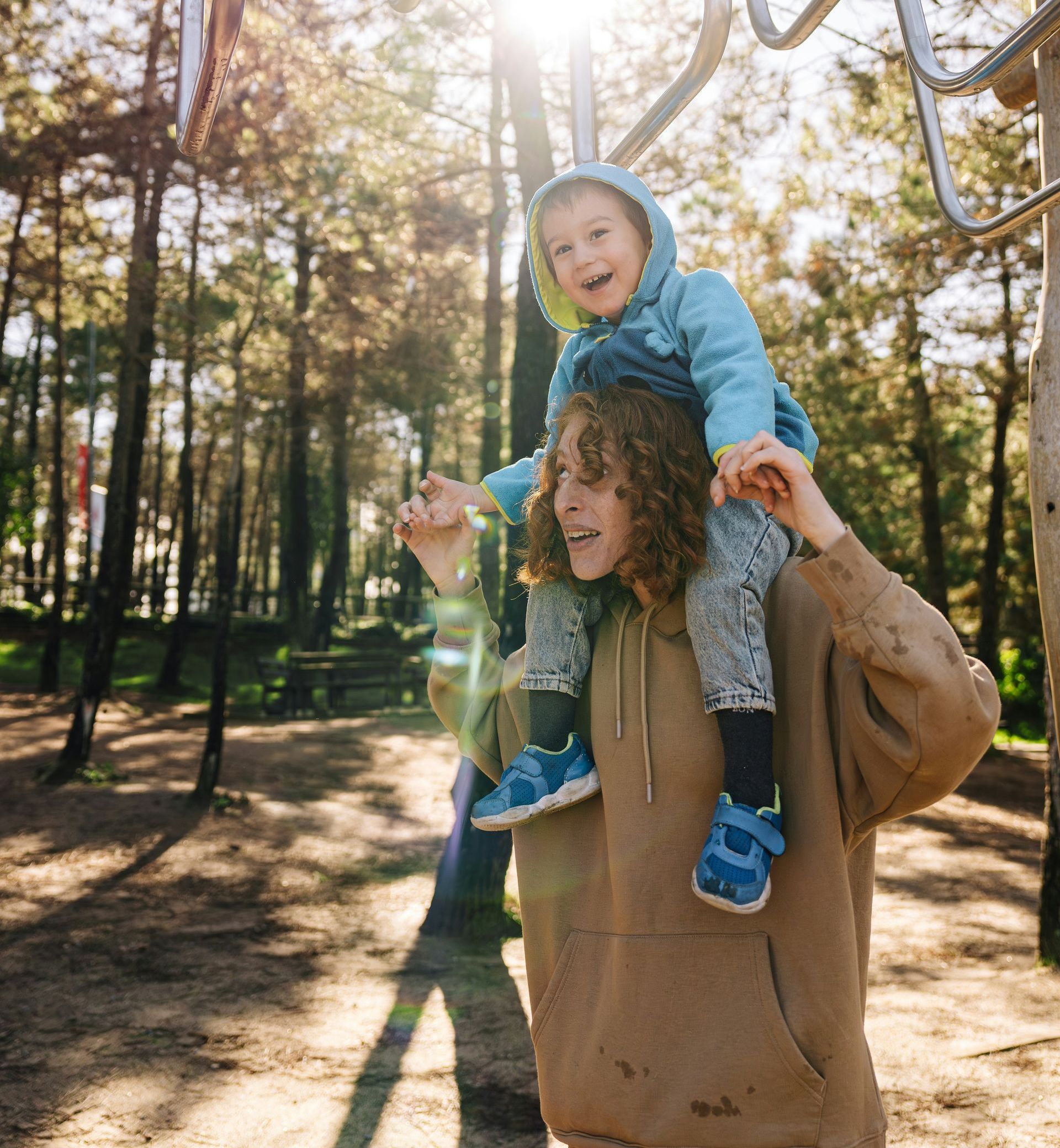 A person in a tan hoodie carries a joyful child in a blue hooded jacket on their shoulders at a sunny, wooded park.