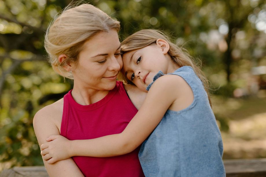 A person in a red top being embraced by a child in a blue top outdoors with a blurred, sunlit background.