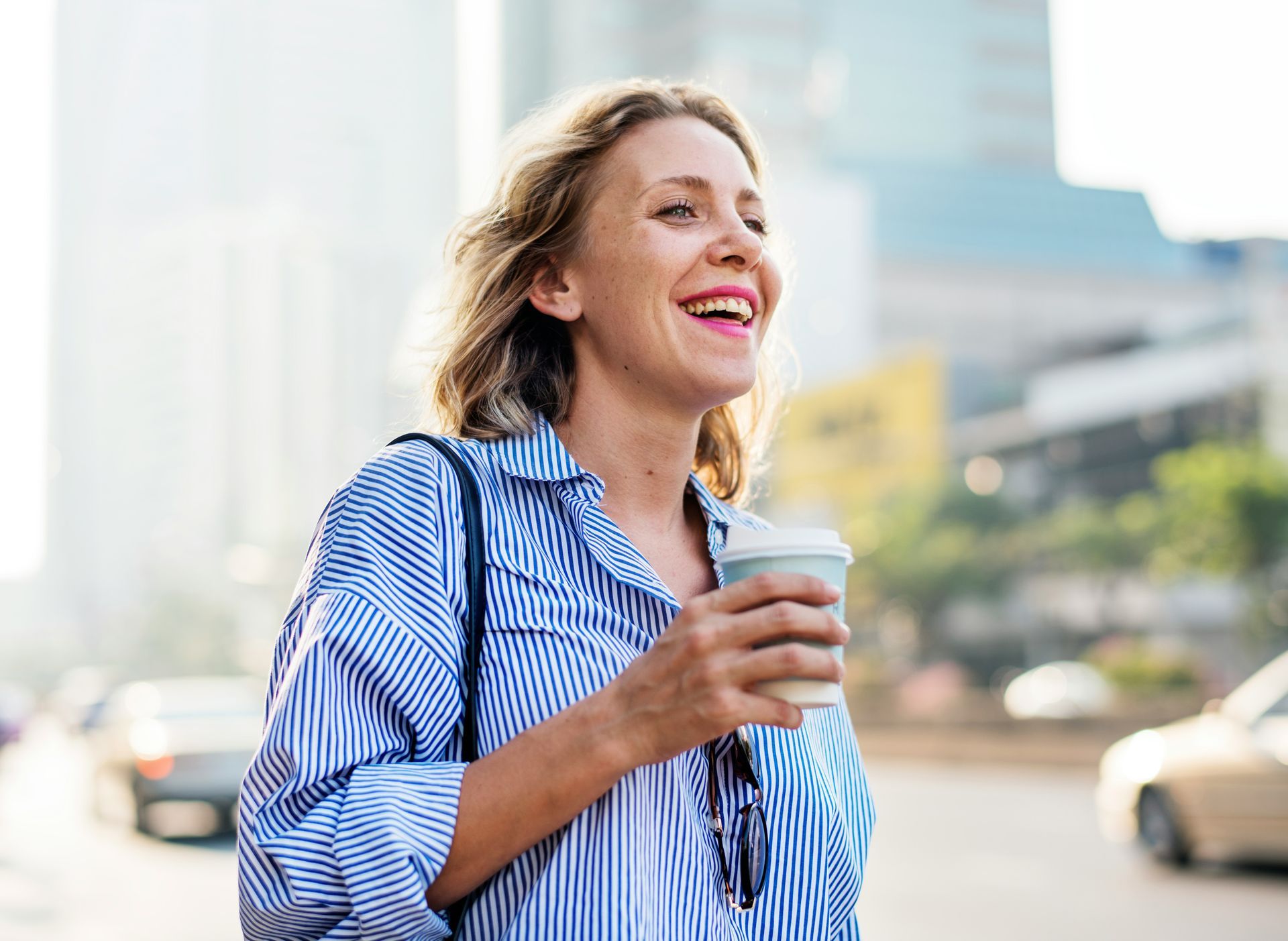 A laughing person in a blue striped shirt walks outdoors, holding a coffee cup in a sunny urban setting.