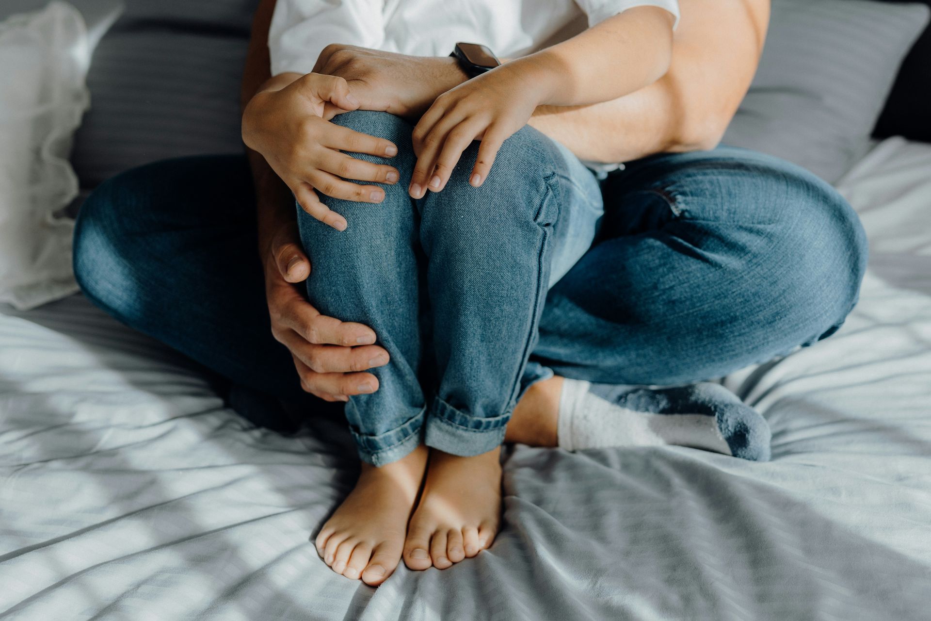 An adult holds a child close, both sitting on a gray bed, with the child’s bare feet resting on the sheets.