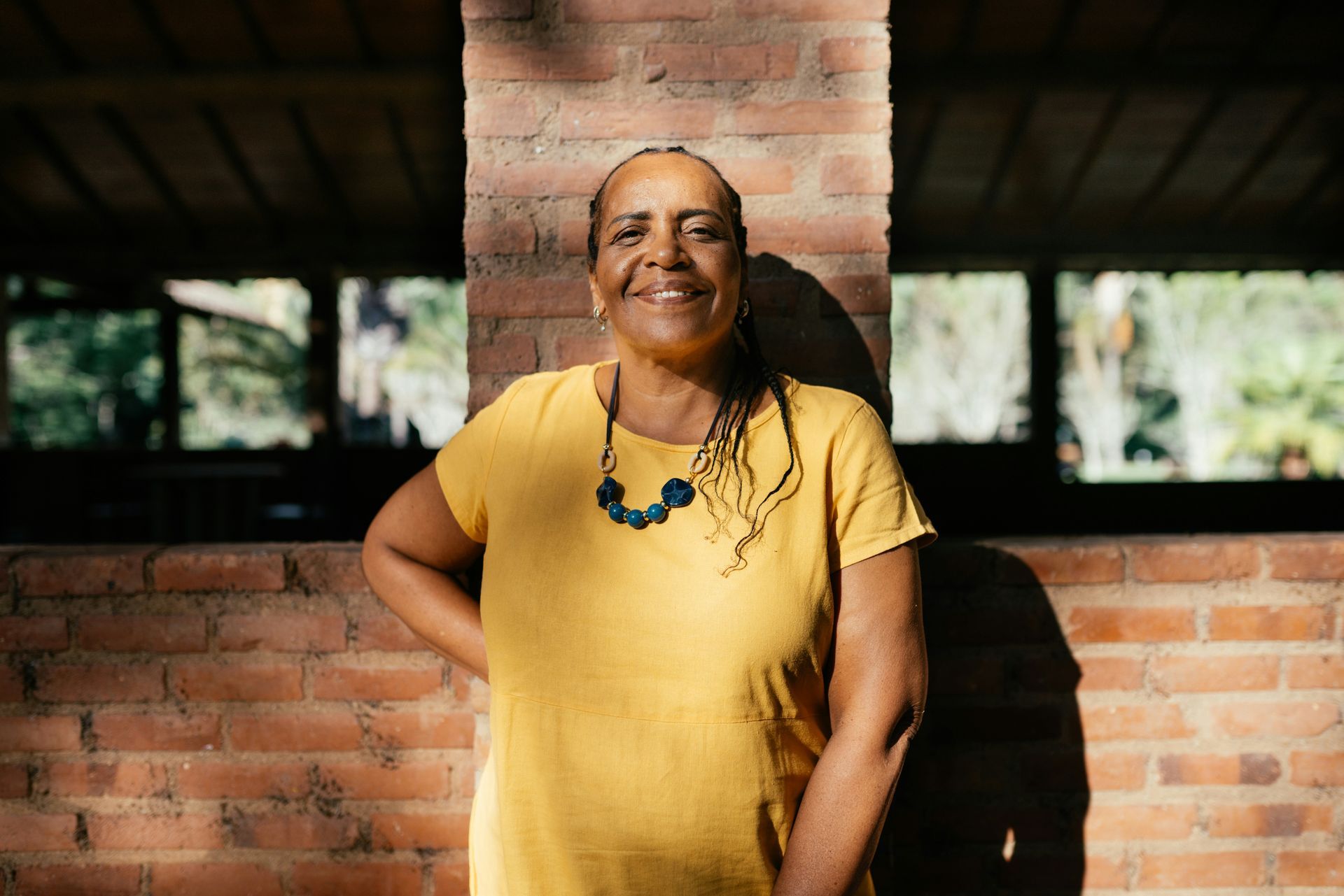 A smiling person in a yellow t-shirt and dark beaded necklace leaning against a brick pillar in an outdoor setting.