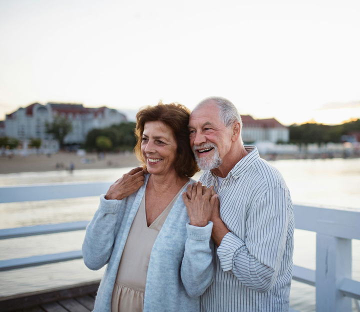 Two people stand closely on a wooden pier by the water, smiling at the horizon with arms around each other.