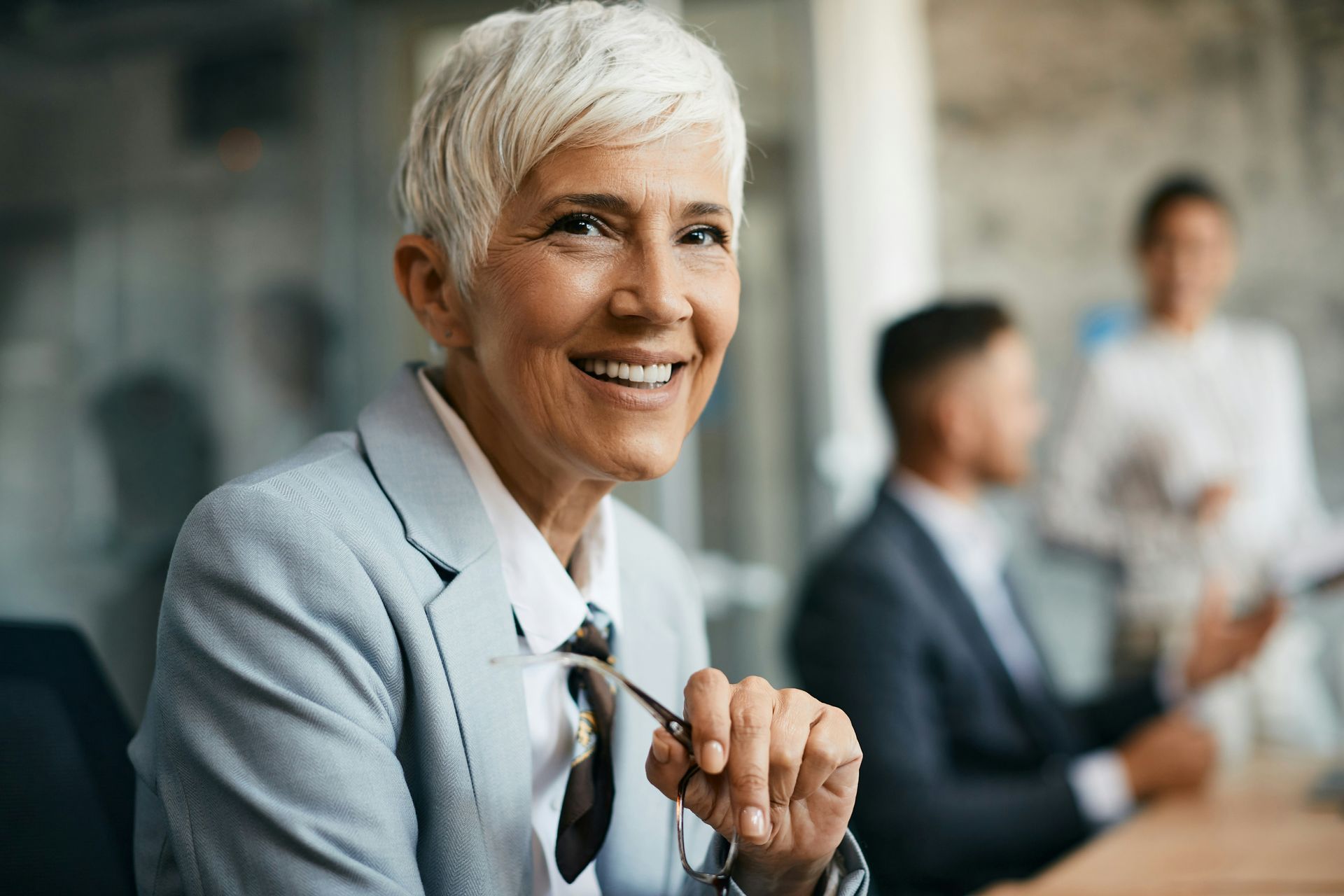 A smiling professional holding glasses in an office, with blurred colleagues working in the background.