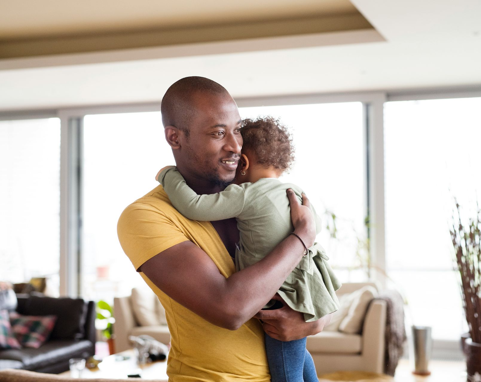 A person in a yellow shirt tenderly holding a small child in their arms in a bright living room.