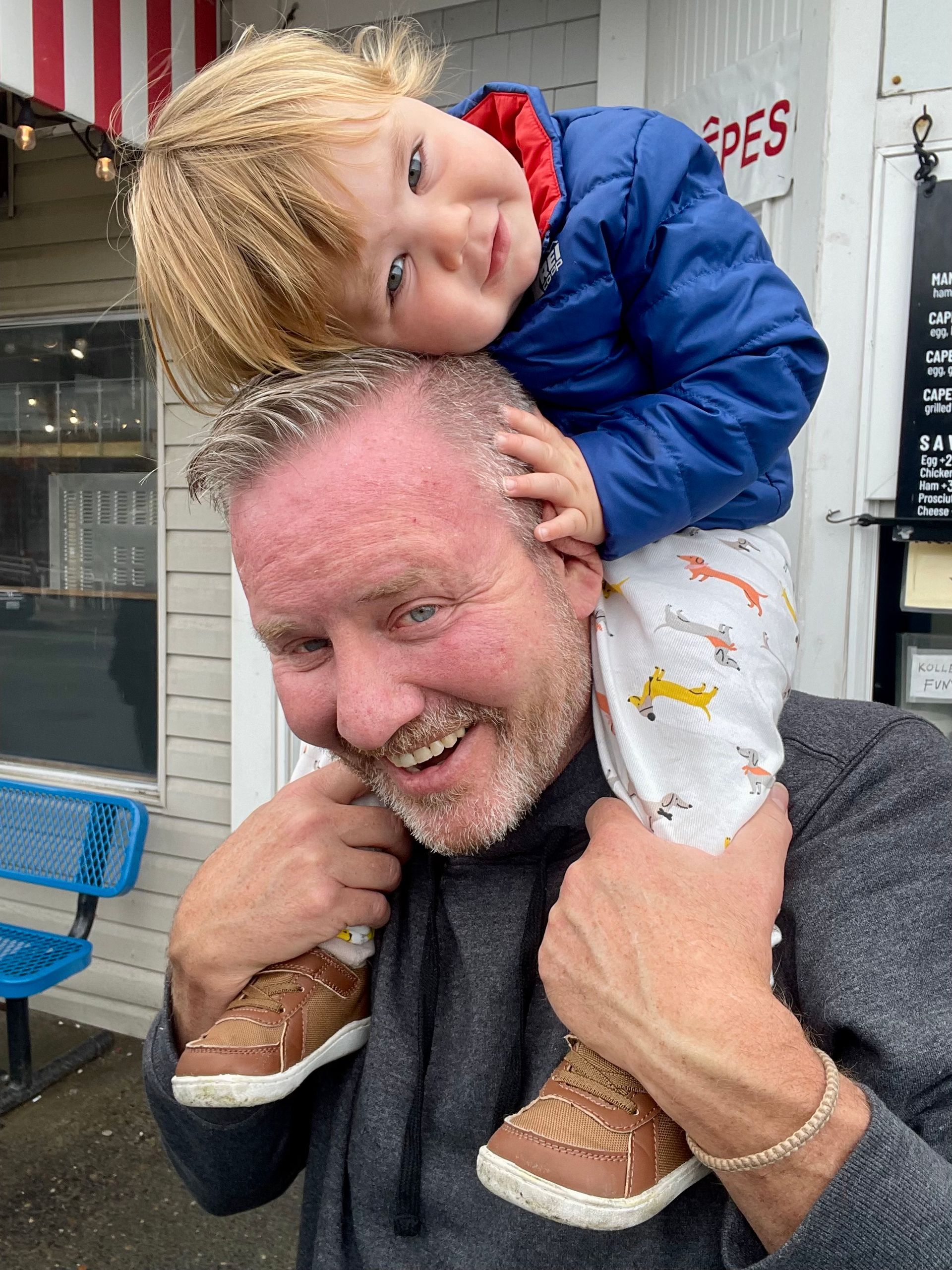 A smiling person carries a young child on their shoulders outside a building.