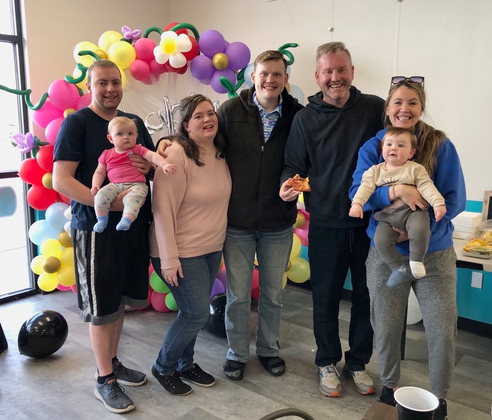 A group of six people stand smiling before a balloon display, two adults holding infants, in an indoor setting.