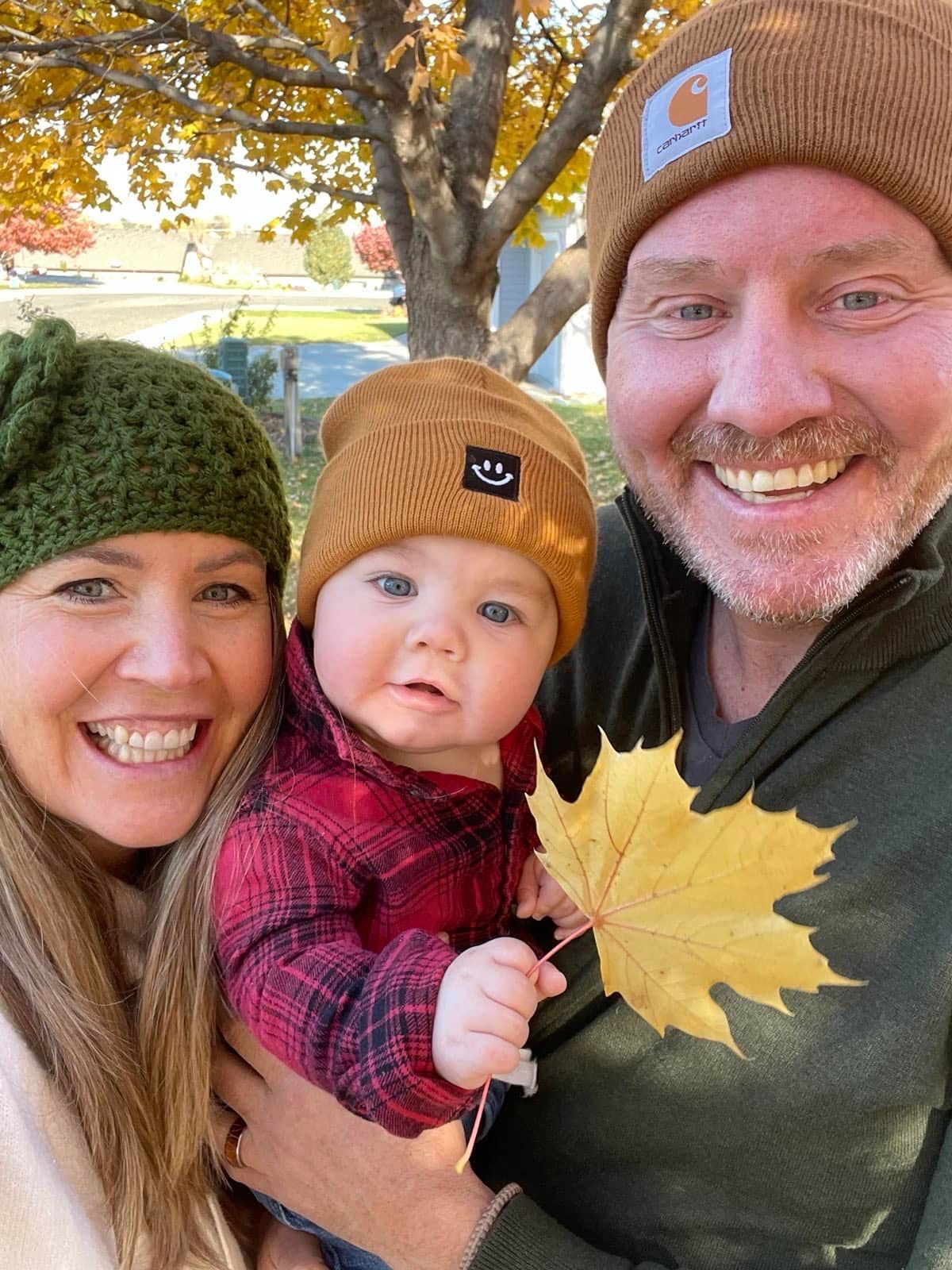 A smiling family wearing beanies poses outdoors in autumn, with the baby holding a yellow maple leaf.