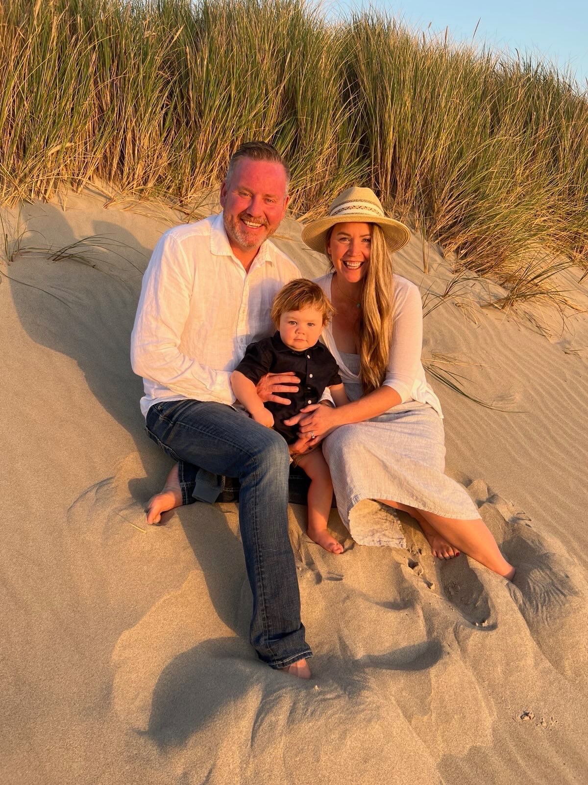 A family of three sitting together on a sandy beach dune during sunset, with tall grass in the background.
