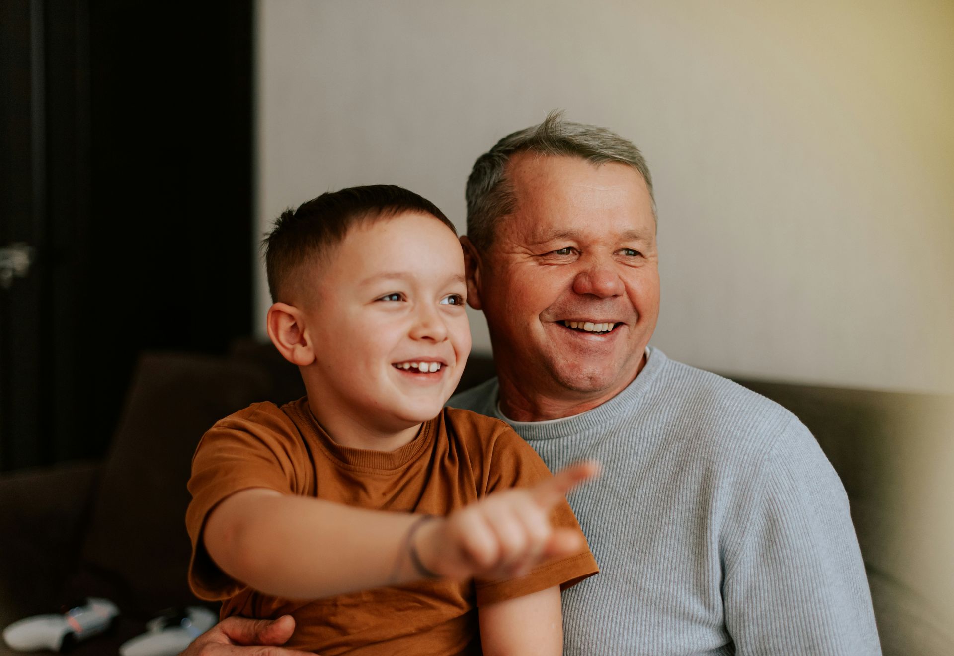 A child in a brown shirt points excitedly while sitting with an adult in a grey sweater, both smiling in a home setting.
