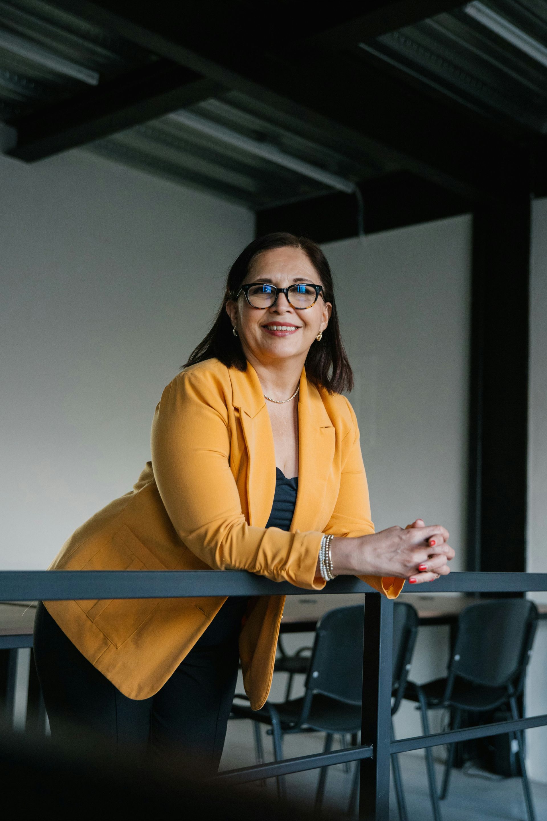 A person in a yellow blazer and glasses leans against a railing in an office, smiling at the camera.