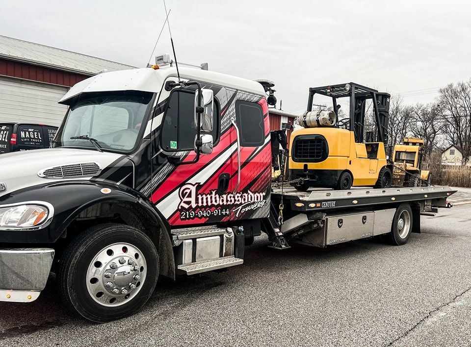 Tow truck with a yellow forklift on its flatbed, parked in front of a building.