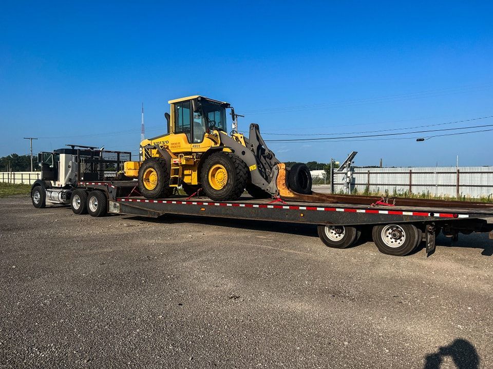 Semi-truck transporting a yellow Volvo wheel loader on a flatbed trailer under a blue sky.