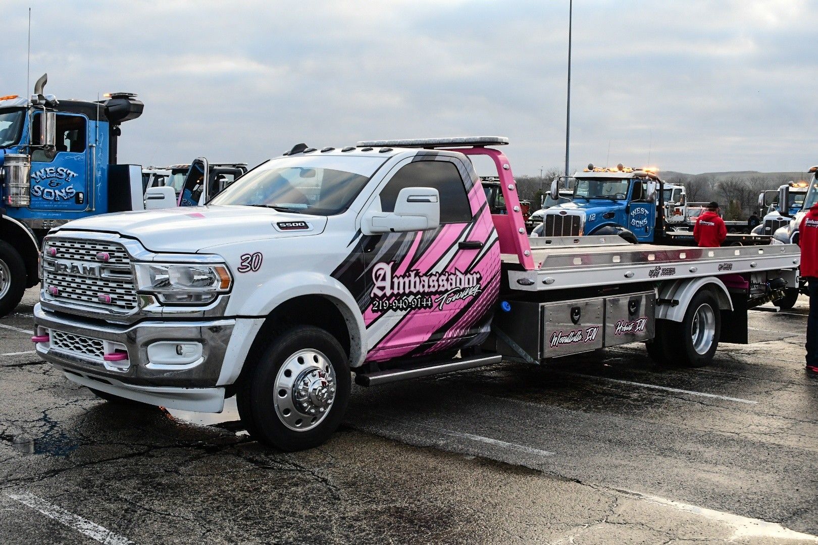 White and pink tow truck parked on asphalt.  Pink graphic on the side, gray sky background.